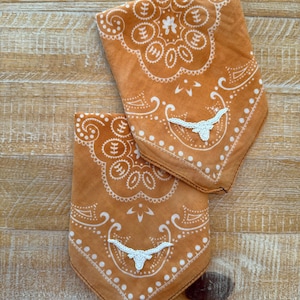 May include: Two rust-colored bandanas with white paisley patterns and a white crocheted longhorn design. The bandanas are folded and displayed on a wooden surface. The bandanas have a classic Western aesthetic.