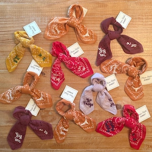 May include: A collection of colorful bandanas in shades of yellow, orange, red, and purple, each tied in a knot. Some bandanas feature embroidered text and designs, with tags that read "Chain Stitch Bandit."