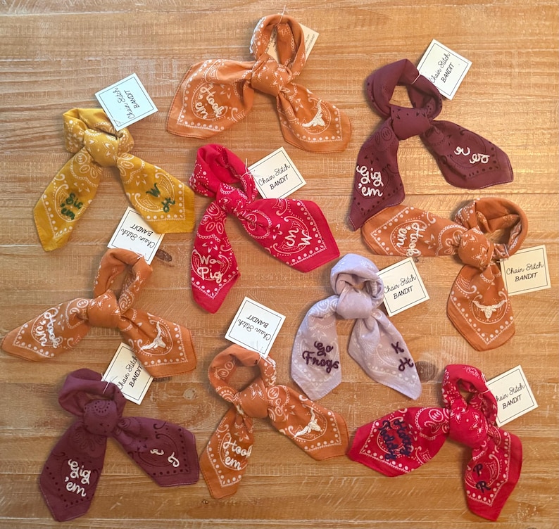 May include: A collection of colorful bandanas in shades of yellow, orange, red, and purple, each tied in a knot. Some bandanas feature embroidered text and designs, with tags that read "Chain Stitch Bandit."