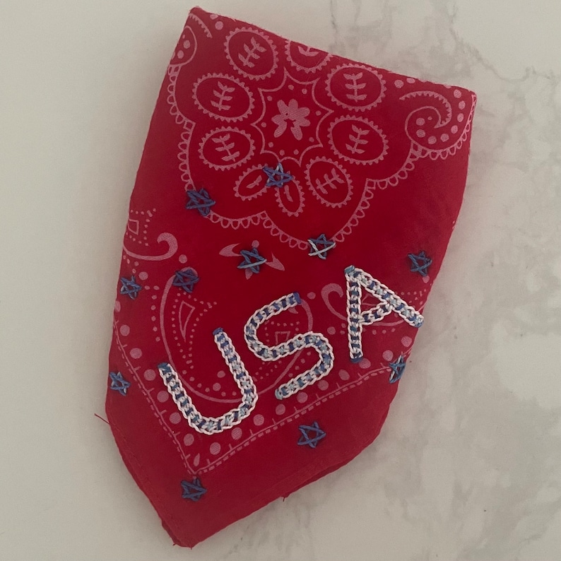 Puede incluir: Bandana roja con un patr&oacute;n de cachemira blanco y estrellas azules. La palabra "USA" est&aacute; bordada en blanco en la bandana.