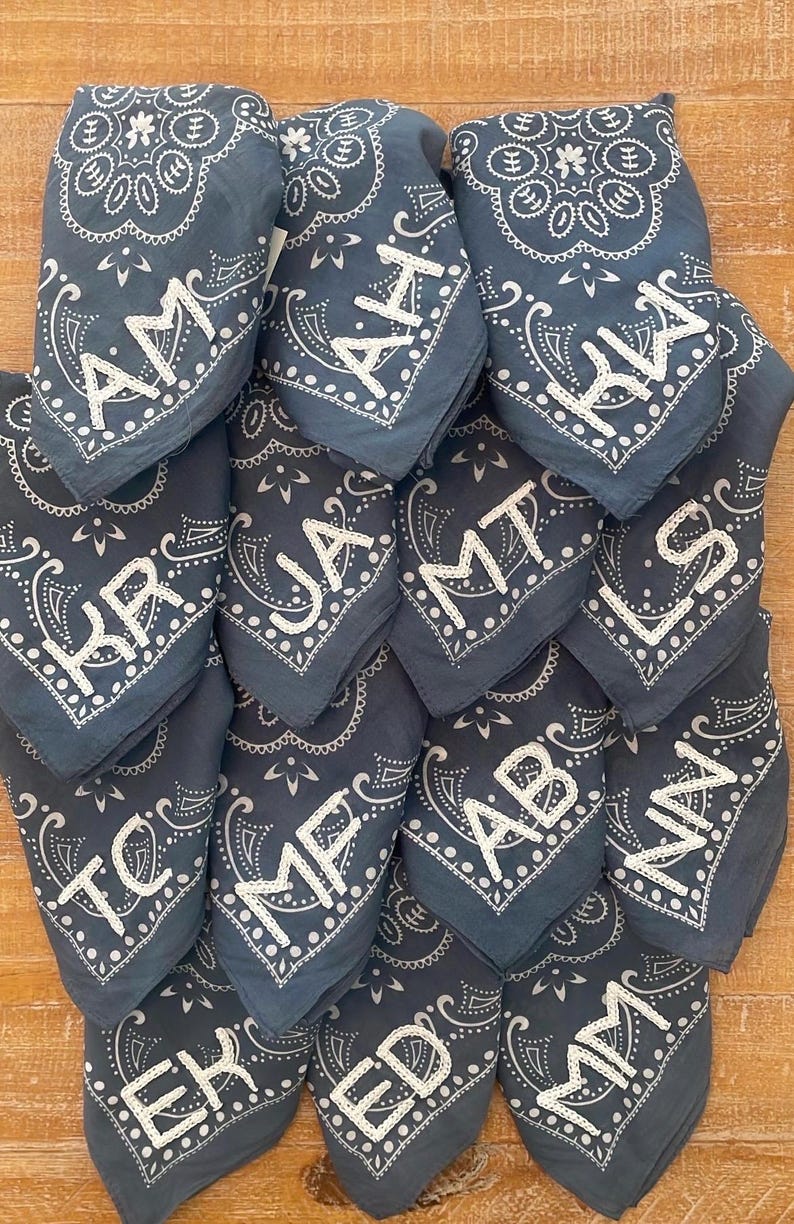 May include: A stack of blue bandanas with white paisley patterns and white embroidered initials. The bandanas are folded in half and stacked on top of each other.
