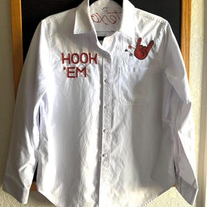 May include: White button-down shirt with red embroidered text that says "HOOK 'EM" on the front. A red hand gesture and stars are embroidered on the right side. The word "Texas" is embroidered on the collar.