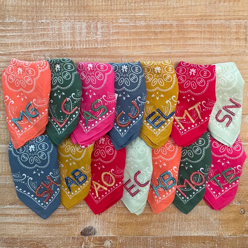 May include: A set of 12 colorful bandanas with embroidered letters. The bandanas are in various colors, including red, blue, green, yellow, and orange. Each bandana has a different letter embroidered on it, from A to L.