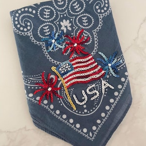 Puede incluir: Un pa&ntilde;uelo azul con un patr&oacute;n paisley blanco. El pa&ntilde;uelo est&aacute; bordado con una bandera estadounidense roja, blanca y azul, un poste amarillo y tres fuegos artificiales rojos, blancos y azules. La palabra "USA" est&aacute; bordada en blanco debajo de la bandera.