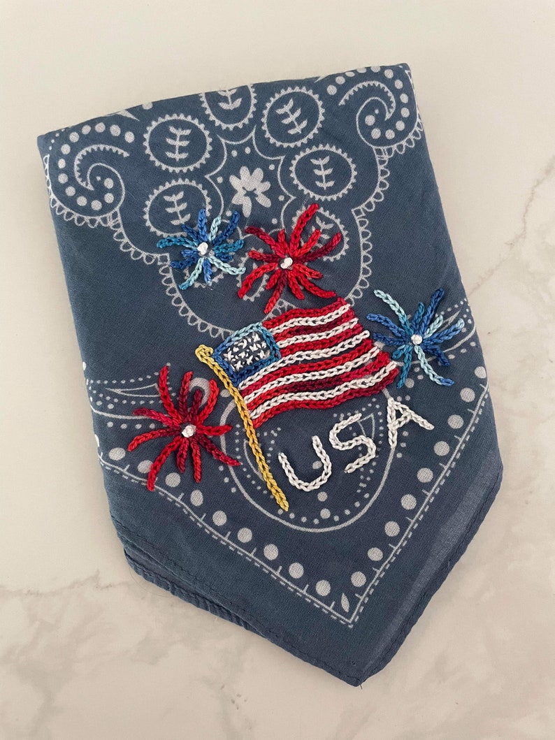 Puede incluir: Un pa&ntilde;uelo azul con un patr&oacute;n paisley blanco. El pa&ntilde;uelo est&aacute; bordado con una bandera estadounidense roja, blanca y azul, un poste amarillo y tres fuegos artificiales rojos, blancos y azules. La palabra "USA" est&aacute; bordada en blanco debajo de la bandera.