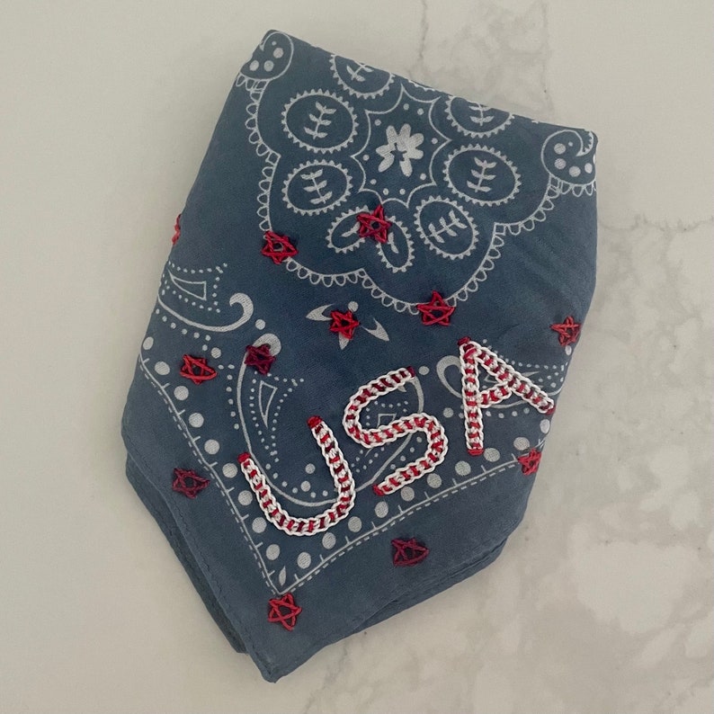 Puede incluir: Un pa&ntilde;uelo azul con un patr&oacute;n paisley blanco y estrellas rojas. La palabra "USA" est&aacute; bordada en blanco con puntadas rojas.