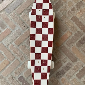 May include: A skateboard with a red and white chequered design. The deck has a striking pattern of alternating red and white squares. The skateboard has white wheels and is designed for recreational use.