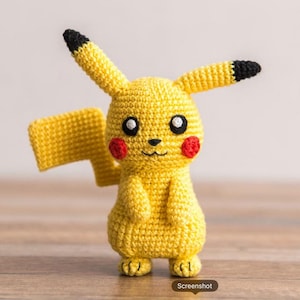 Puede incluir: Un peluche de ganchillo amarillo que representa un personaje con orejas largas de puntas negras, mejillas rojas circulares y una cola en forma de rayo. La figura tiene ojos negros con reflejos blancos y una pequeña boca bordada.