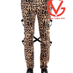 May include: Leopard print trousers featuring black and tan spots, a button fly, and black straps with silver-toned metal rings attached to the sides. The trousers are paired with black and white zebra-striped shoes. A red logo with the text Gothic Vibes NYC.