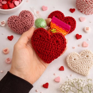 Könnte beinhalten: Eine Hand hält ein rotes gehäkeltes Herz, mit einem kleinen Bonbon und einem Reese's. Verstreut sind gehäkelte Herzen in Rosa und Creme, kleine Holzherzen und Herzbonbons. Das Thema ist der Valentinstag.