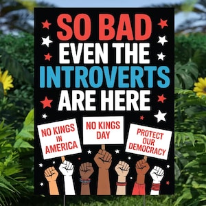 May include: A black yard sign with the text "SO BAD EVEN THE INTROVERTS ARE HERE." Below, signs read "NO KINGS IN AMERICA," "NO KINGS DAY," and "PROTECT OUR DEMOCRACY." The sign is supported by a metal stake and set in a grassy area.