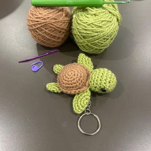 May include: A crocheted turtle keyring with a brown shell and green body, beside two balls of yarn, a green crochet hook, a purple yarn needle, and a purple stitch marker. The turtle has a silver keyring.
