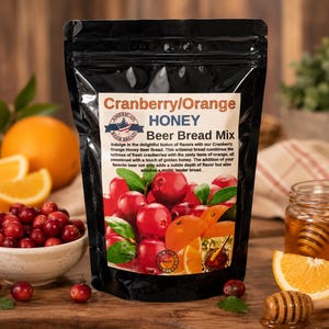May include: A black stand-up pouch containing Cranberry/Orange Honey Beer Bread Mix. The label features text, a circular logo with a flag, and a graphic of fresh red cranberries and orange slices. The pouch is sealed with a top zipper closure.