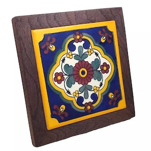 May include: Square decorative tile with a floral design in a dark wood frame. The tile features a blue background with a central floral motif in red, white, and green, surrounded by yellow and red accents. A decorative piece for home decor.