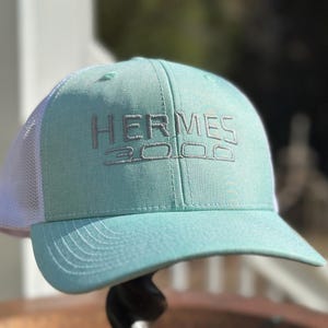 May include: A turquoise and white trucker hat with the text "HERMES 3000" embroidered in silver on the front.