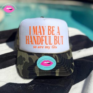 Puede incluir: Gorra de camionero blanca y camuflada con el texto "I MAY BE A HANDFUL BUT so are my tits" en naranja. La gorra tiene una visera curva y una parte trasera de malla. Una pegatina con labios rosas y las palabras "funny mother truckers".