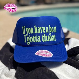 May include: A blue trucker hat with the text "If you have a boat I gotta throat" in green. The hat has a mesh back and a curved brim. A sticker with pink lips and the words "Funny Mother Truckers" is on the front.