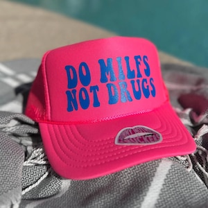 以下が含まれることがあります： 鮮やかなピンク色のトラッカーハット。青い文字で「DO MILFS NOT DRUGS」と書かれています。メッシュの背面と湾曲したつばが付いています。唇のステッカーがつばに貼られています。