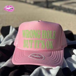 May include: Pink trucker hat with the text "WRONG HOLE BUT IT'S OK" in green. The hat has a mesh back and a curved brim. A small sticker with the text "FUNNY MOTHER SUCKERS" is on the side.