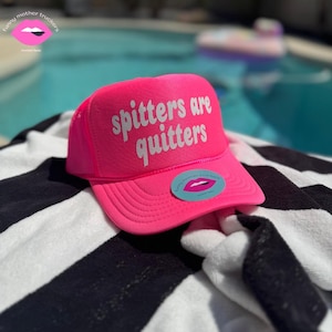 May include: Pink trucker hat with a white mesh back. The hat has the text "Spitters are quitters" in white letters on the front.