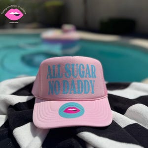 Puede incluir: Gorra de camionero rosa con una malla azul en la parte posterior y el texto "ALL SUGAR NO DADDY" en letras azules. La gorra tiene una pequeña pegatina rosa con un labio rosa y el texto "funny mother truckers" en ella.