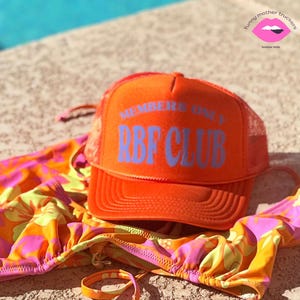 Könnte beinhalten: Orangefarbene Trucker-Kappe mit weißem Netzrücken. Die Kappe trägt den Schriftzug "Members Only RBF Club" in blauer Schrift.