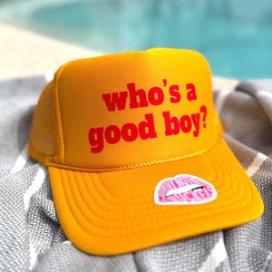 Puede incluir: Gorra de camionero de color amarillo mostaza con la frase "who's a good boy?" en letras rojas. Una pegatina de labios rosas y blancos está en la visera. El sombrero está sobre una toalla a rayas grises y blancas.