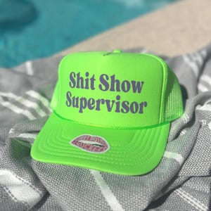 May include: Bright neon green trucker hat with the text "Shit Show Supervisor" in purple. The hat features a mesh back and a curved brim. A small sticker of lips is on the front.