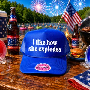 May include: A blue trucker hat with the text "i like how she explodes" in white. The hat is on a wooden surface with a bottle of beer, a hamburger, and an American flag in the background. The hat has a "Tiny Mouth Truckers" sticker.