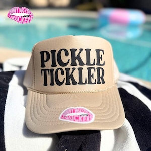 Op de afbeelding: Beige truckerpet met de tekst "PICKLE TICKLER" in zwarte letters. Een roze lippensticker met de tekst "FUNNY MOTHER TRUCKERS" staat op de voorkant. De pet ligt op een zwart-wit gestreepte handdoek.