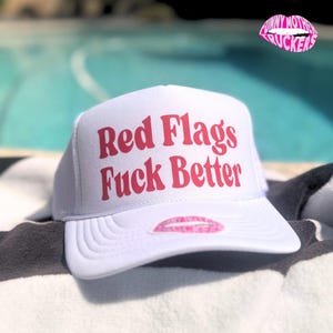 Könnte beinhalten: Weiße Trucker-Mütze mit dem Text "Red Flags Fuck Better" in Rot. Die Mütze hat eine rosa Lippen-Grafik auf dem Schirm und ein "Funny Mother Truckers"-Logo. Die Mütze liegt auf einem Handtuch in der Nähe eines Pools.