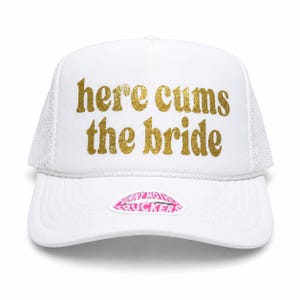 May include: A white trucker hat featuring a mesh back and a white foam front panel. The text "here cums the bride" is printed in a gold glitter font across the front. A small pink and white sticker with the text "FUNNY MOTHER TRUCKERS" is on the brim.