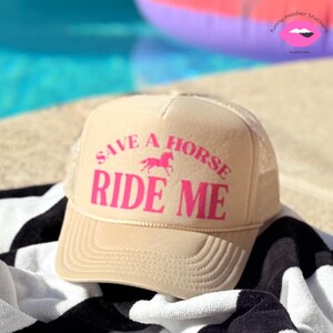 Puede incluir: Una gorra de camionero beige con un gráfico de caballo rosa y el texto "Save a Horse Ride Me" en rosa.