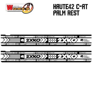 以下が含まれることがあります： 「HAUTE42 C-AT PALM REST」のテキストが入った、白と黒のパームレスト。デザインには、「2XKO」と「RIOT GAMES」のロゴ、矢印と幾何学模様が含まれています。「O.D.M. WORKS」のロゴも表示されています。