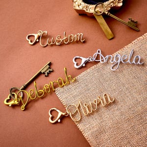 May include: A collection of custom metal name keychains featuring cursive script lettering. The set includes gold-toned wire names Deborah and Olivia, a silver-toned wire name Angela, and a gold-toned wire word Custom, each attached to a heart-shaped clasp.