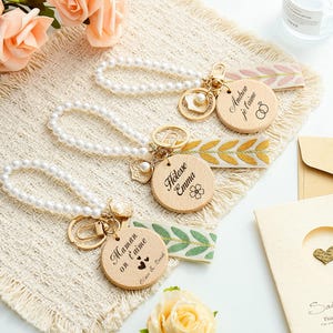 May include: Three keychains featuring white pearl loops, gold-tone metal clasps, and fabric ribbons with leaf patterns. Each includes a round wooden disc engraved with text: Andrew je t'aime, Hôtesse Emma, and Maman on t'aime with heart icons.