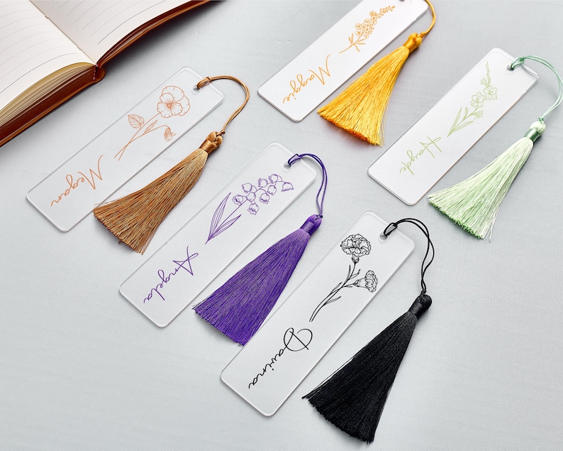 Op de afbeelding: Transparante acryl bladwijzers met bloemenillustraties en gepersonaliseerde namen. Elke bladwijzer heeft een bijpassende kwast. De namen zijn Megan, Maggie, Hayle, Angela en Davina. De kwasten zijn bruin, geel, groen, paars en zwart.