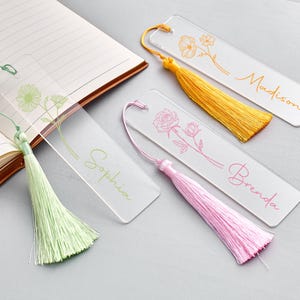 Puede incluir: Marcadores de acrílico transparente con diseños florales y nombres personalizados. Cada marcador presenta una borla de diferente color: verde, amarillo y rosa. Los nombres "Sophia", "Madison" y "Brenda" están escritos en cursiva.