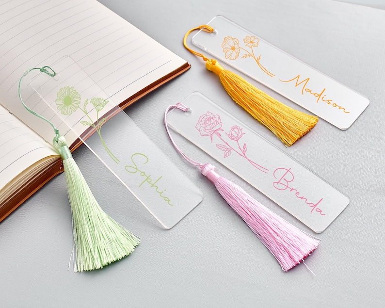 Op de afbeelding: Transparante acryl bladwijzers met bloemmotieven en gepersonaliseerde namen. Elke bladwijzer heeft een andere kleur kwast: groen, geel en roze. De namen "Sophia", "Madison" en "Brenda" zijn in cursief geschreven.