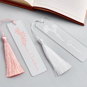 Op de afbeelding: Drie transparante acryl bladwijzers met bloemmotieven en kwastjes. E&eacute;n bladwijzer heeft een roze kwast en de naam "Carolina" in bijpassend roze schrift. Een andere heeft een witte kwast en de naam "Shannon" in witte letters. Een open boek staat op de achtergrond.