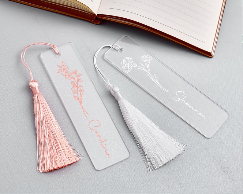 Op de afbeelding: Drie transparante acryl bladwijzers met bloemmotieven en kwastjes. E&eacute;n bladwijzer heeft een roze kwast en de naam "Carolina" in bijpassend roze schrift. Een andere heeft een witte kwast en de naam "Shannon" in witte letters. Een open boek staat op de achtergrond.