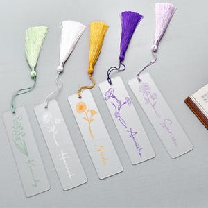 Puede incluir: Una colección de marcadores de acrílico transparente con borlas de colores. Cada marcador presenta un diseño floral y un nombre en fuente cursiva. Las borlas son en tonos verdes, blancos, dorados, morados y rosas. Un cuaderno es visible en el fondo.