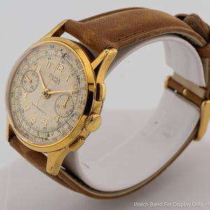 May include: A gold-tone chronograph wristwatch featuring a cream-coloured dial with gold Arabic numerals, two sub-dials, and a tachymeter scale. The watch face displays the text OLMA and 17 RUBIS. It is attached to a brown leather strap.