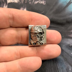 May include: A silver-colored metal money clip featuring a detailed relief of a Stormtrooper helmet. The clip is rectangular with a slightly curved design, showcasing the helmet's iconic features.
