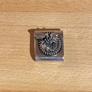 May include: A silver money clip with a detailed, circular, raised design. The design features a central figure surrounded by intricate details. The clip is rectangular with a curved edge for holding bills.