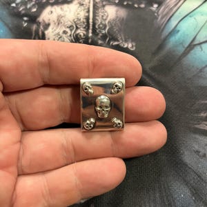 Könnte beinhalten: Ein kleiner, silberfarbener Metallanhänger mit rechteckiger Form. Der Anhänger zeigt einen zentralen Totenkopf und vier kleinere Totenköpfe an jeder Ecke. Das Design ist detailliert und im gotischen Stil gehalten.