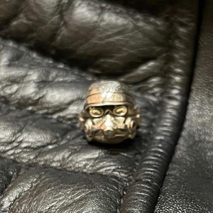 May include: A silver-colored ring shaped like a Stormtrooper helmet. The ring has detailed features, including the helmet's visor and breathing apparatus. The ring is set against a dark, textured background, possibly leather.