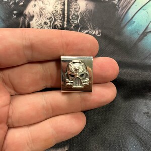 May include: A silver-colored metal rectangular clip featuring a detailed, embossed design of a helmeted face. The face has intricate details, including a visor and facial features. The clip is held in a hand against a patterned background.