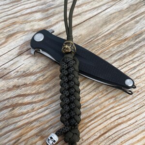 May include: A black folding knife with a green paracord lanyard. The lanyard features a decorative brass bead and a small white skull charm. The knife and lanyard rest on a wooden surface.