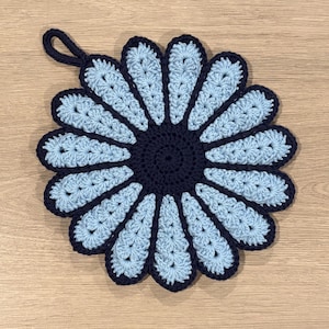 Könnte beinhalten: Ein handgefertigter Topflappen in Blumenform aus Häkelarbeit. Er hat eine dunkelblaue Mitte und Blütenblattumrisse mit hellblauen Blütenblättern. Eine Schlaufe zum Aufhängen ist angebracht. Der Topflappen hat einen Durchmesser von ca. 20 cm.